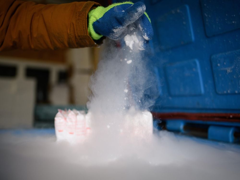 Someone wearing a thick blue glove drops dry ice on a surface at the bottom of the frame, causing white fog to spread 