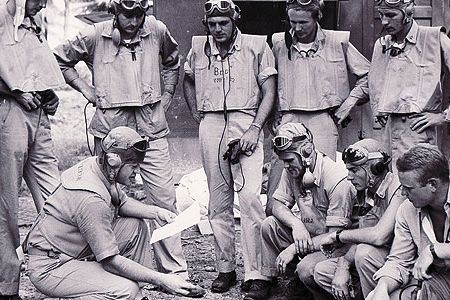 At the Black Sheep Squadron's base on the South Pacific island of Espiritu Santo, Boyington (holding paper) briefs his pilots on an upcoming mission.