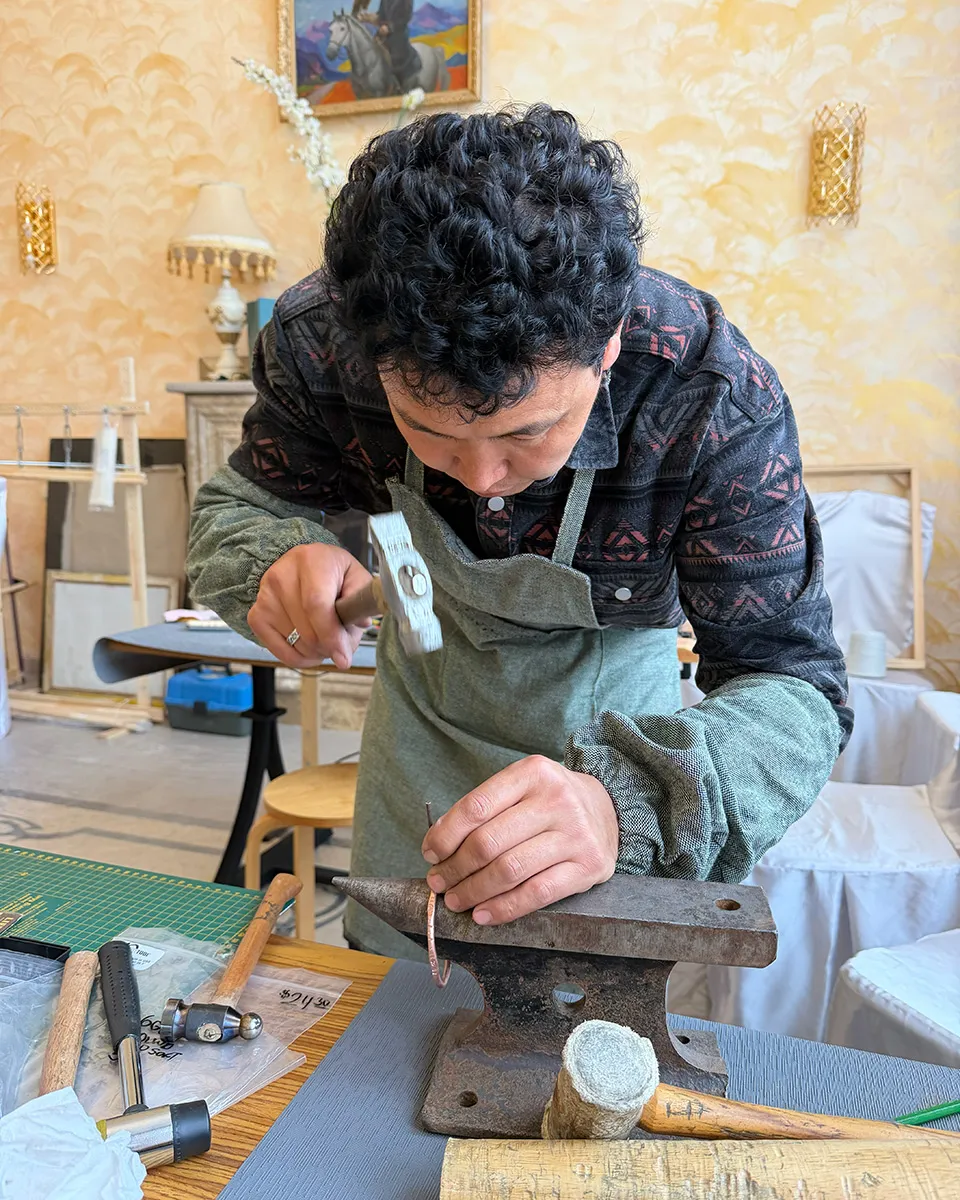 A man holds a copper wire to an anvil with one hand and prepares to strike it with a hammer with the other.