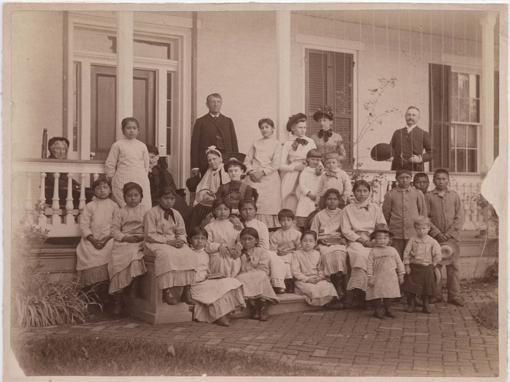 A sepia toned image of a group of young women, with a few young boys and girls, sitting in front of a white house on a porch. Pratt stands in dark garb behind the group