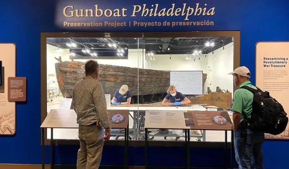 Visitors look into an exhibit window and see staff working on the wooden Gunboat