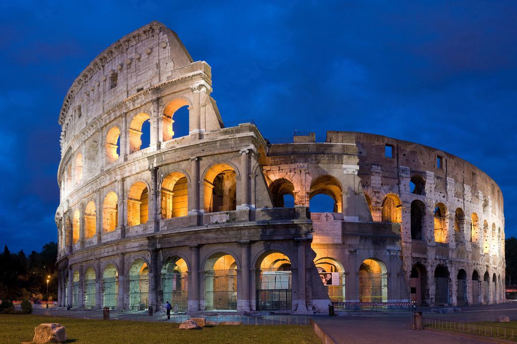 The Colosseum at night, with a blue sky and its arches lit up by yellow light. The structure is partially dismantled, with half its walls torn down
