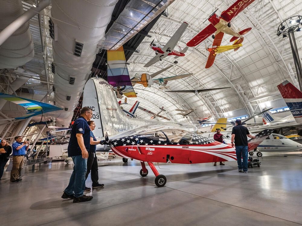 Inside a large hangar filled with natural light, three white people maneuver a small red, white, and blue airplane into its display space. At least eight aircraft are suspended from the hangar's ceiling.