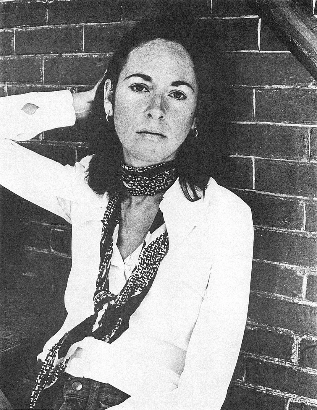 A young, elegant Gluck leans against a brick wall in a black and white image; holds one hand up behind her head in a pose, looks seriously at camera; in a white shirt with shoulder length dark hair, earrings, and a thin scarf tied loosely around her neck