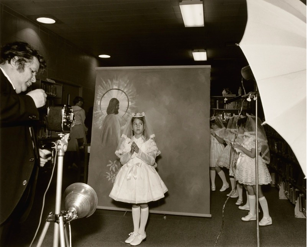 Black and white photograph of a girls posing for a first communion picture. A dropdown background image of a man with a halo around his head. A photographer stands with a camera in front of the girl.