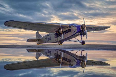 A vintage silver Ford Tri-Motor aircraft parked on a wet tarmac under a dramatic, cloudy sky at dusk. The plane features corrugated metal skin, three engines, and the "Ford" script logo in blue on the fuselage, all reflected in the water on the ground.