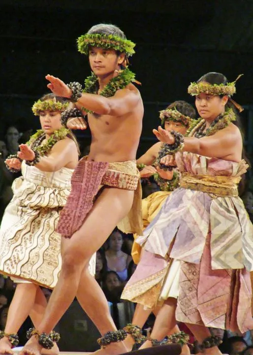 Four hula dancers perform on stage, wearing green leis and crowns and pastel-colored bark cloth dresses or loin cloths.