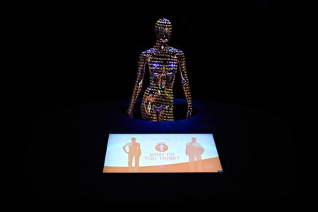 A wide shot of a museum exhibit, featuring a dark, faceless, naked female mannequin covered in orange genetic code in the background with a monitor in the foreground showing an orange and white graphic of two people with "What do you think?" between them.