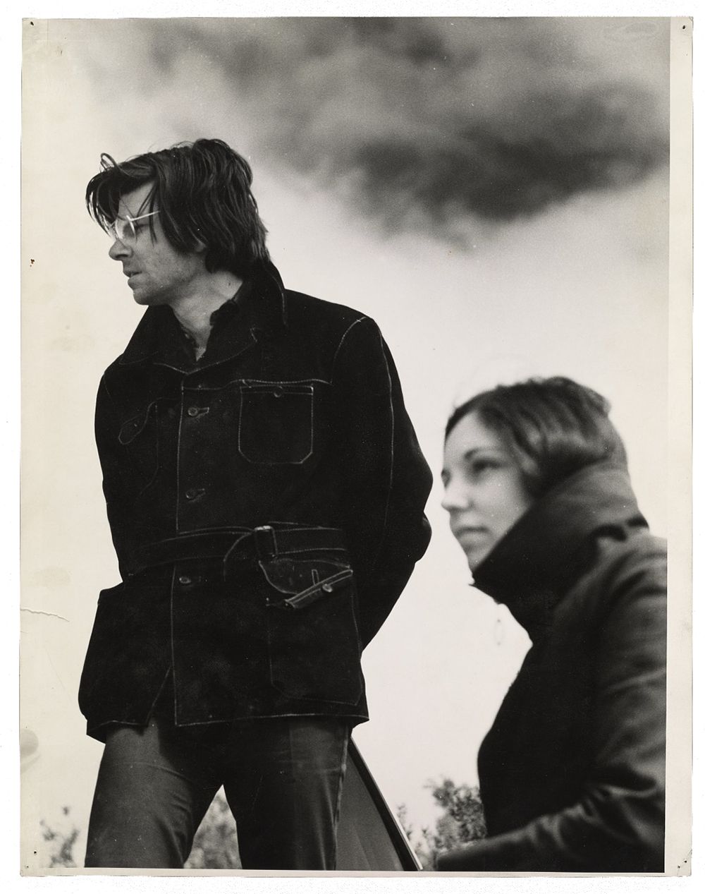 Grayscale image showing Nancy Holt out of focus in the right corner with Robert Smithson in focus in the main frame. Both are looking off into the distance in coats with expressionless faces.