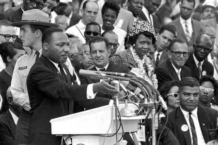 Martin Luther King, Jr. stands at a podium with several microphones. Behind him to his left is a crowd of seated and standing people, including Dorothy Height, turned towards him.