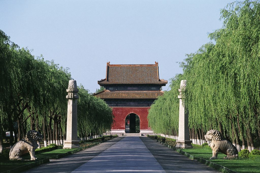 Red building at the end of a long road lined with trees and stone statues