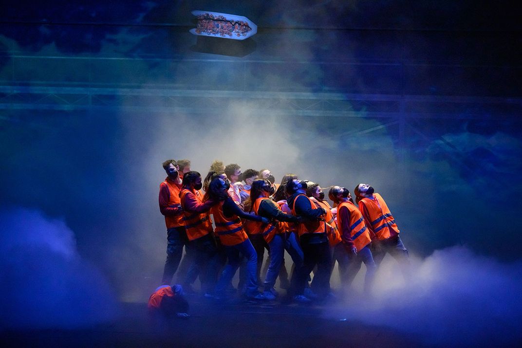 Scene on a stage of several people huddled togethered in orange life vests. Fake fog and eery blue light surrounds them.