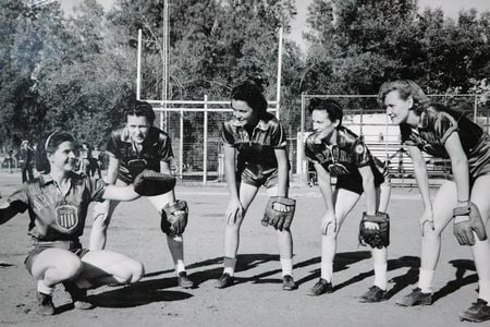 A group photo of women in softball uniforms wearing baseball gloves pose for a photo