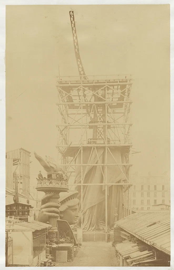 Assemblage of the Statue of Liberty in Paris, showing the bottom half of the statue erect under scaffolding, the head and torch at its feet, at the Gaget, Gauthier & Co. workshop in Paris, 1881-1883