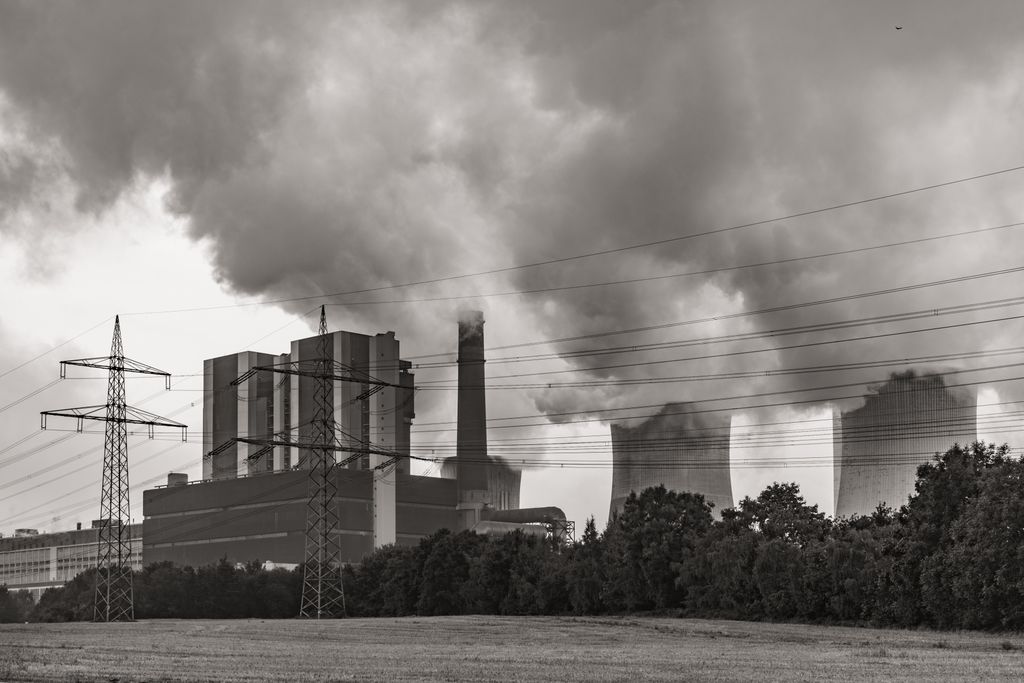 In black and white, power lines cross the foreground as a power plant billows smoke behind it.