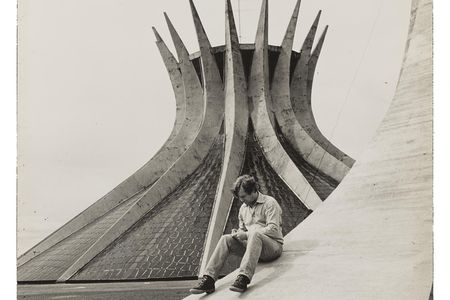 Grayscale image of Robert Hughes sitting on the sloping curve of a building examining something in his hands, with a modernist church in the background.