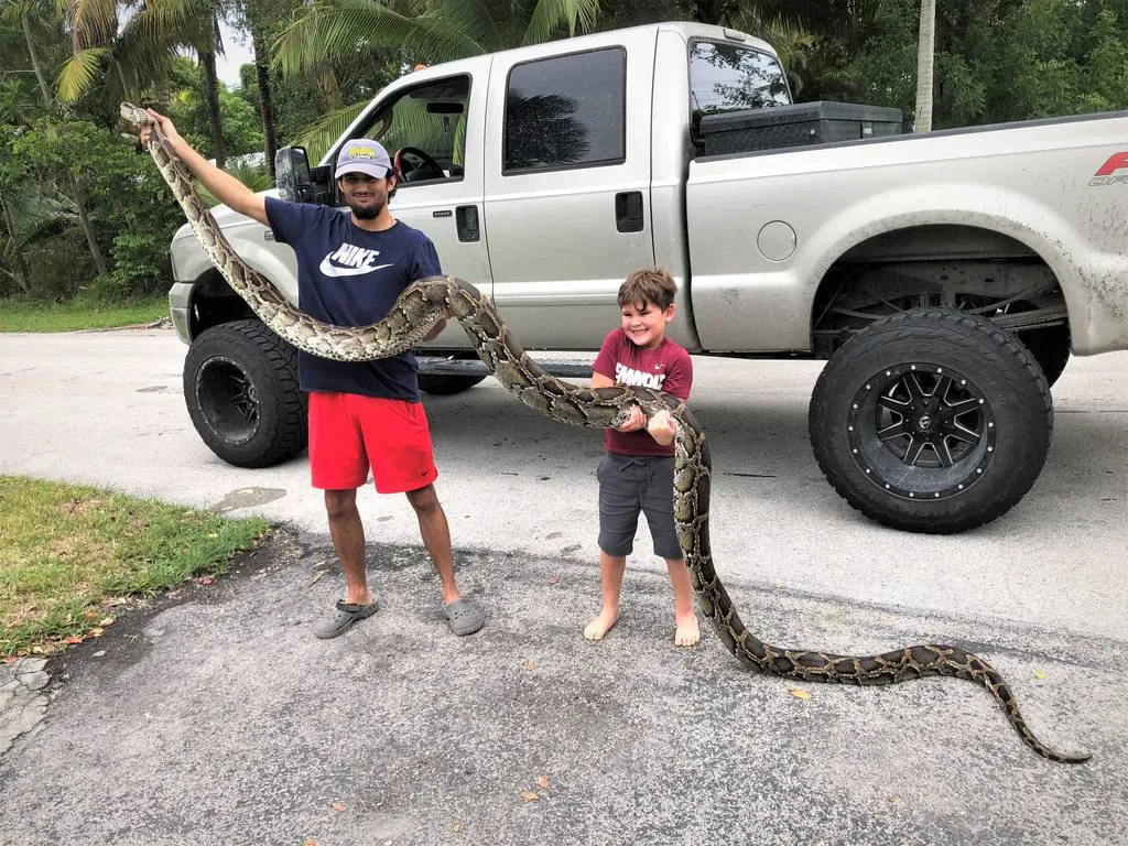 Two kids holding a long snake with a truck in the background