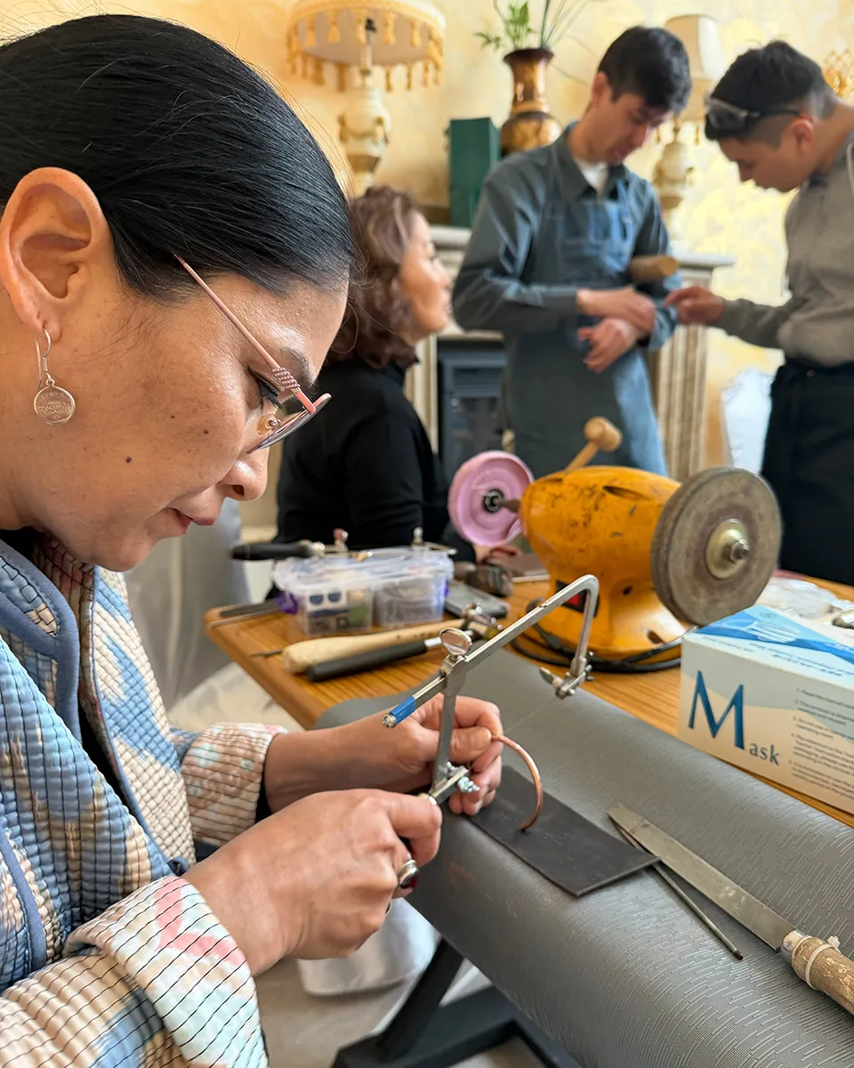 A woman with black hair and glasses concentrates on using a metal tool to shape a copper bracelet. Two others behind her examine a bracelet too.