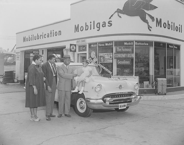 A car salesman and family stand outside a gas station