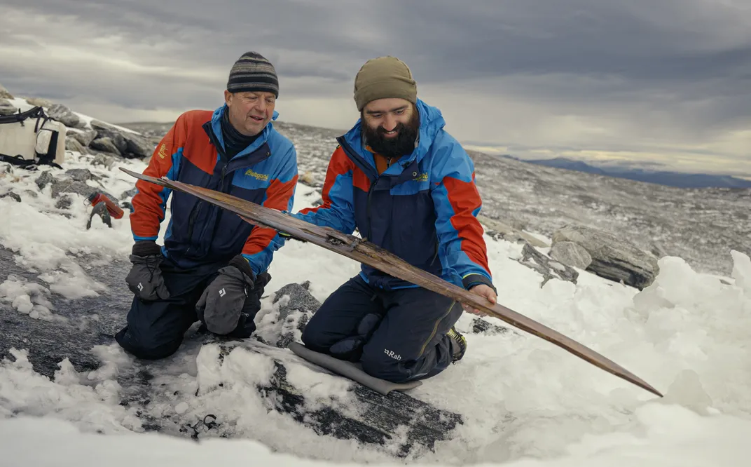 two men kneel on the ice, one holding a long double-pointed stick