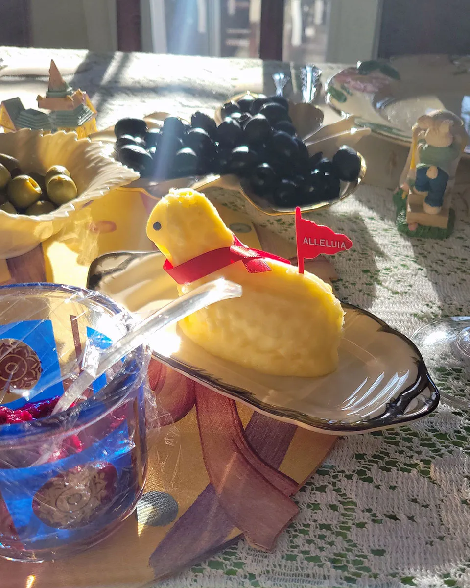 A festive table setting featuring a butter shaped like a lamb surrounded by olives, nuts, and decorations under natural sunlight.