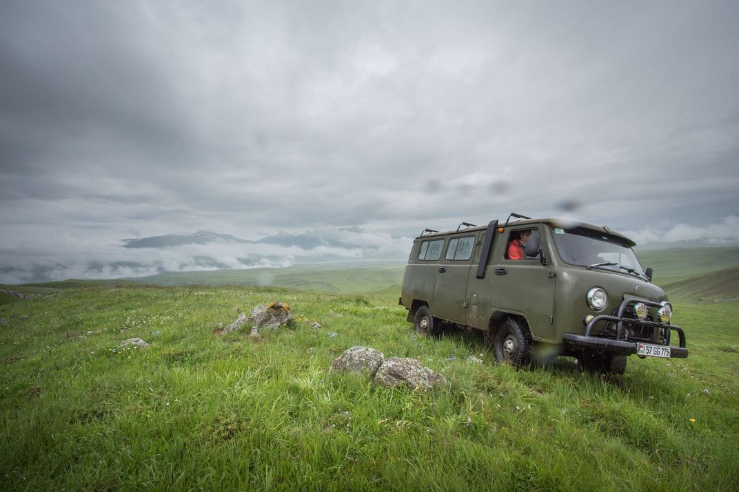 A vintage, dark green van drives through a misty field atop a mountain.