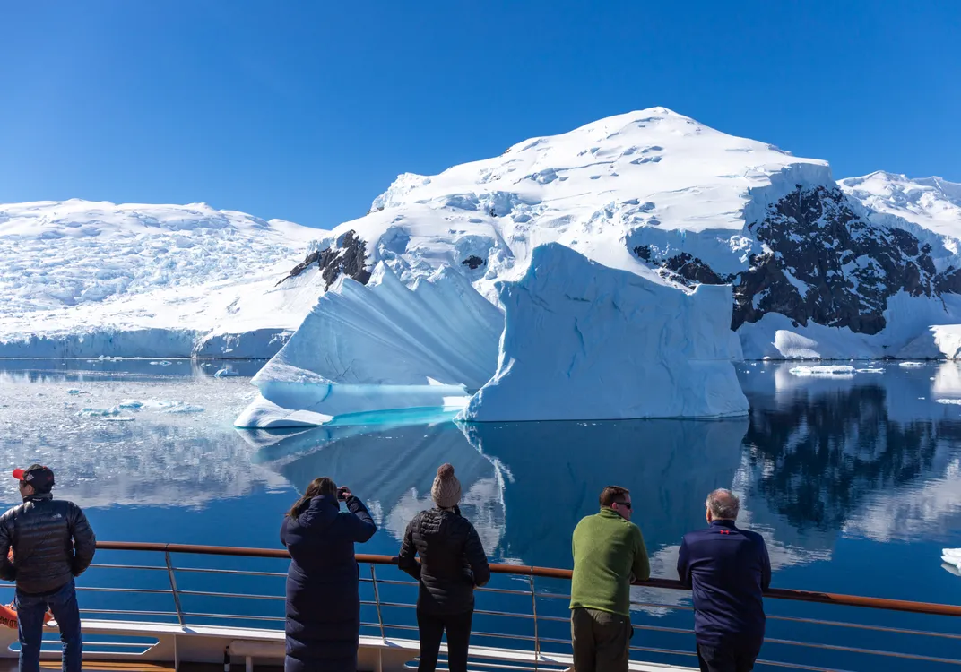 Four people stand on the deck of a ship, looking at a massive iceberg and snow-covered mountains reflected in the calm water of an Antarctic landscape.