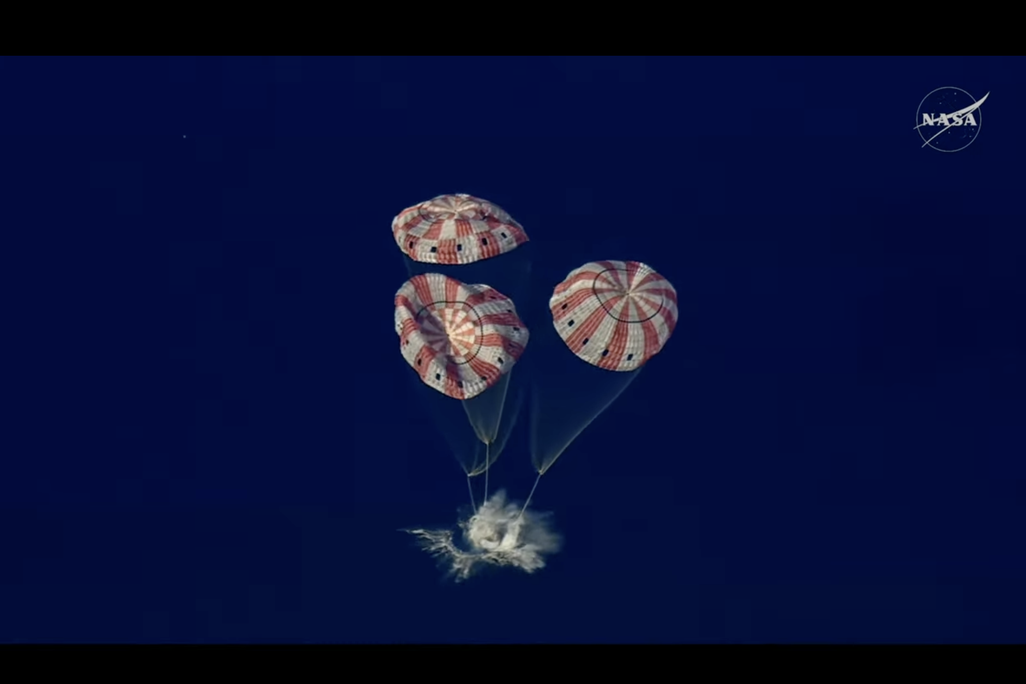 spacecraft splashing down in the ocean with three parachutes above