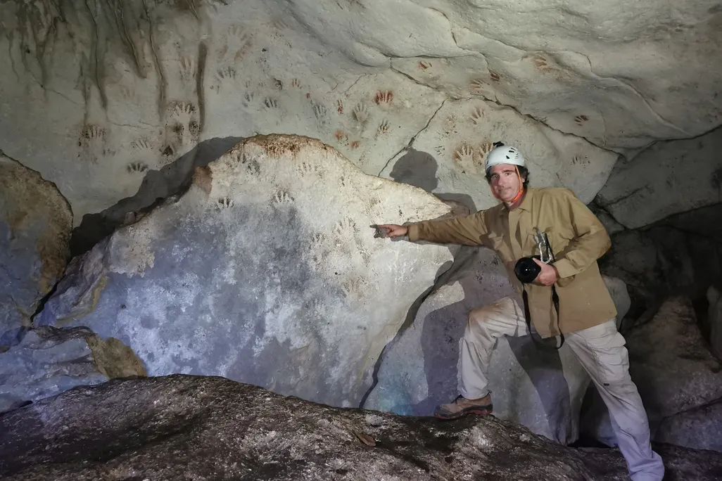 A man wearing boots and a white helmet stands in front of a large rock and points to handprints, which are reddish or white against black shadows and cover the sloping wall of the cave