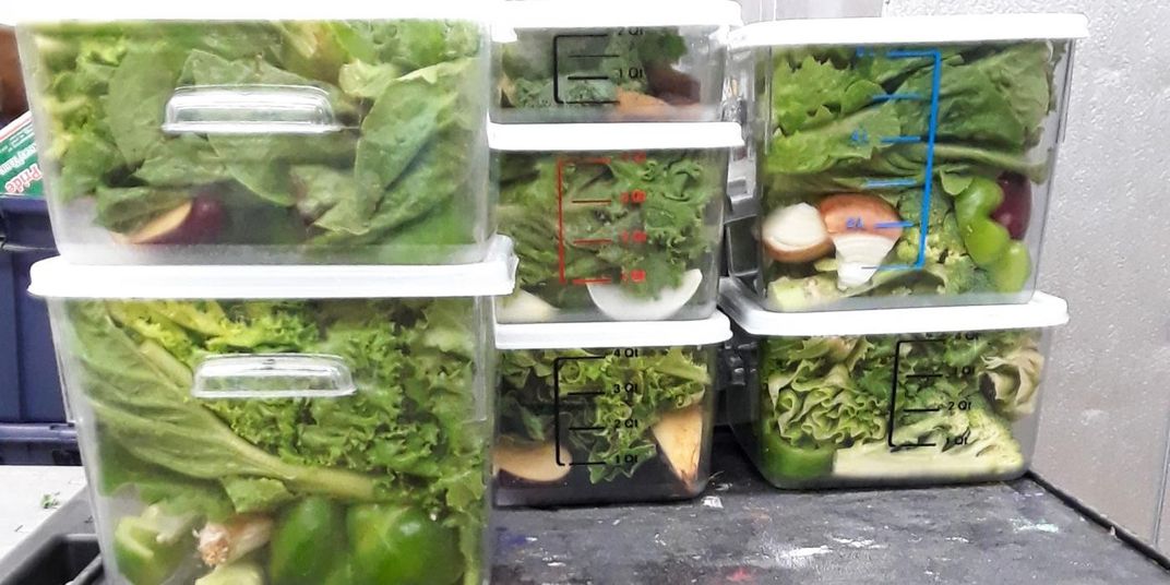 Plastic bins full of lettuce and chopped fruits and vegetables stacked on a table.