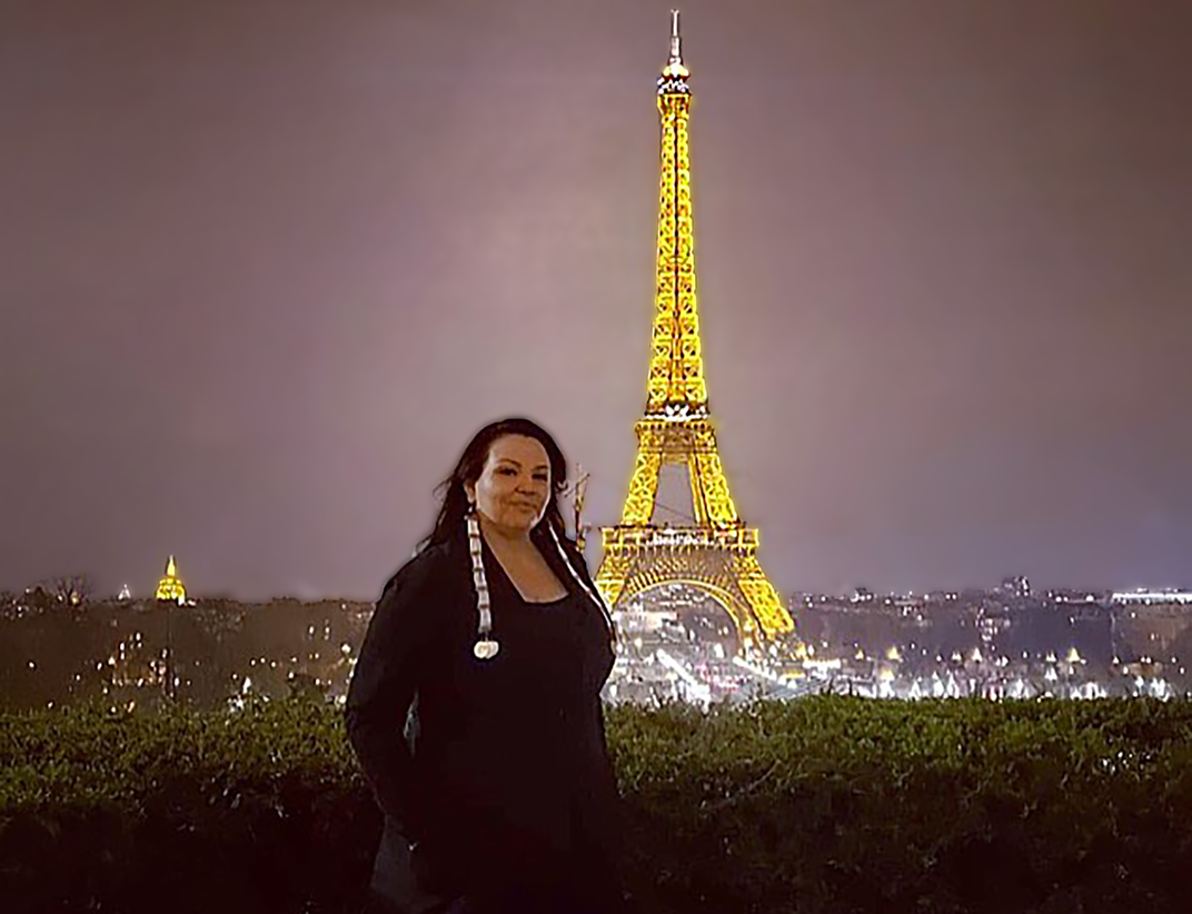 The fashion designer Norma Baker–Flying Horse, dressed in a black gown and gloves, stands outdoors in Paris at night. The Eiffel Tower is lit in the far background.