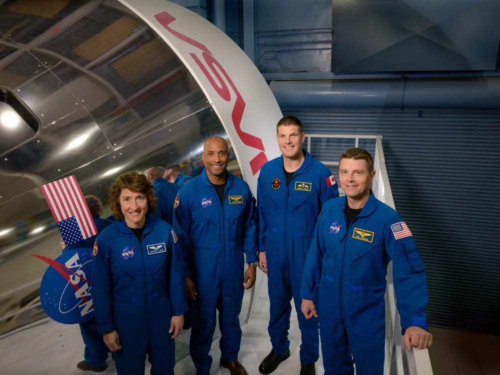 Four astronauts stand smiling in blue suits