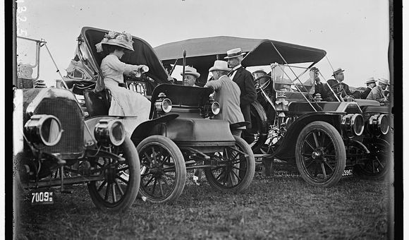 A black and white photograph featuring cars with people conversing.
