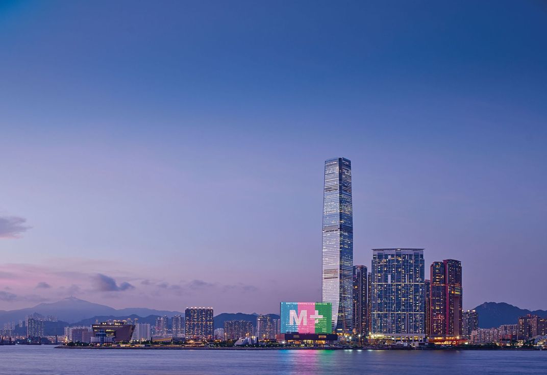 A dramatic shot of the M+ museum's illuminated facade on the waterfront, in front of Hong Kong's skyline and a blue and purple twilit sky