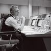 A black-and-white image of a white man in his early 40s sitting at a work console with two display screens and dozens of buttons. The man is wearing a short-sleeve shirt covered by a black vest.
