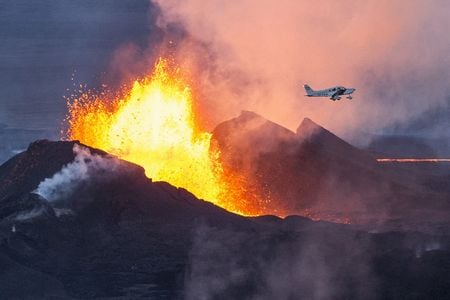 Red hot molten lava and thick dark dust spew from the ground while a small white. single-propeller aircraft flies overhead.