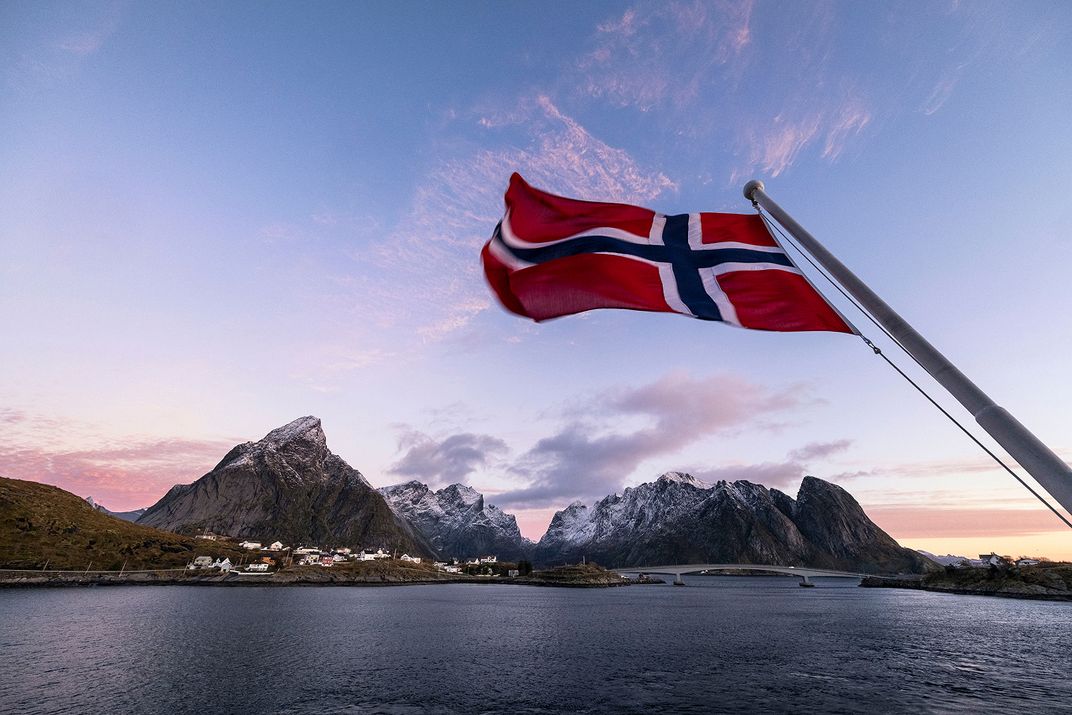 A Norwegian flag flies above coastal waters in the Lofoten Islands, with rugged snow-capped mountains, a bridge, and small seaside villages visible beneath a colorful sunset sky.