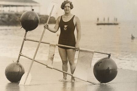 A black and white gelatin silver print of Gertrude Ederle standing on the shore with a 1920s flotation device.