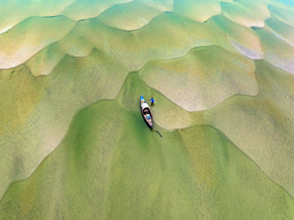 In a drying river among Bangladesh’s chars, a lone boat sits pinned between rippled sandbanks and shallow green water, its tiny fisherman revealing how quickly flowing lifelines can turn into fragile, shifting ground.