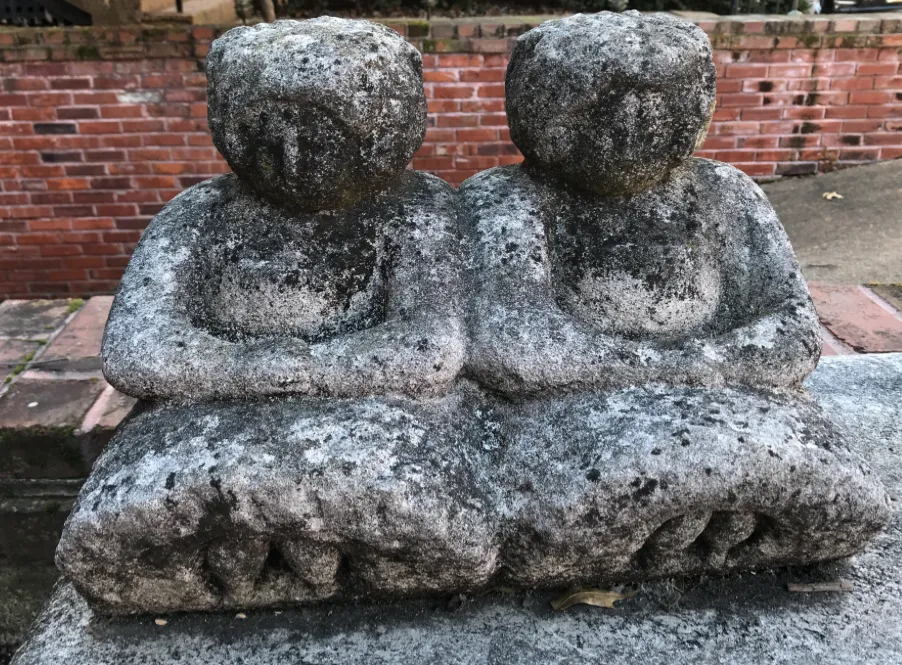 A sculpture of two seated women, covered in moss, dirt and weathered with time, rests outside in front of a sidewalk and brick wall
