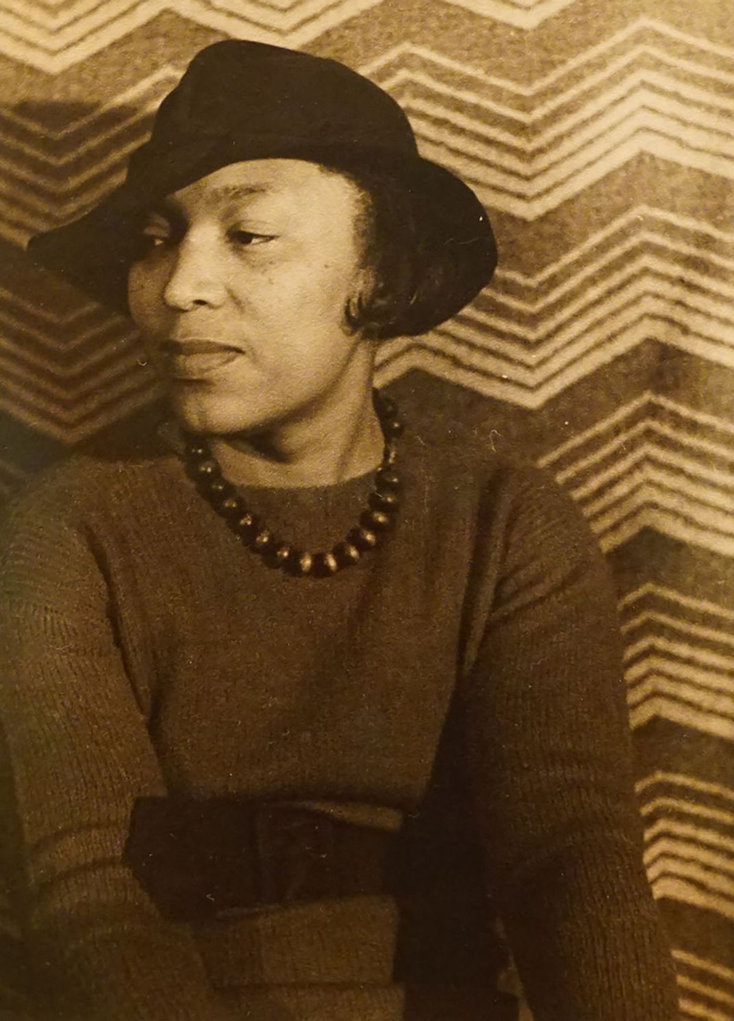 portrait of Zora Neale Hurston wearing a dark hat in front of a pattered cloth backdrop