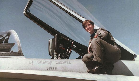 A young woman wearing a U.S. Navy olive-green flightsuit and black combat boots perches next to the cockpit of her F/A-18C miltary jet. "Lt Sharon Cummins" is stenciled onto the side of the airplane.