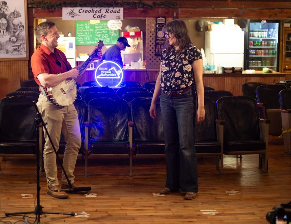 A man stands indoors while holding a banjo and smiling at a woman. The woman stands and smiles back at the man.