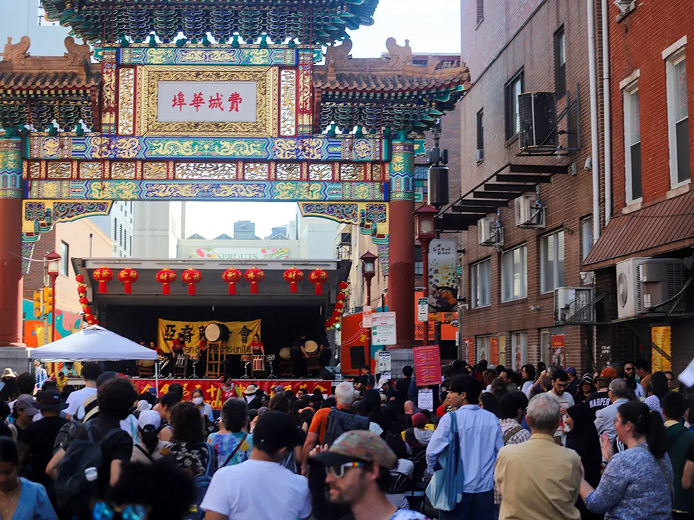 A crowd gathers on a street, where a stage is set up under an elaborate, decorative Chinese-style archway.