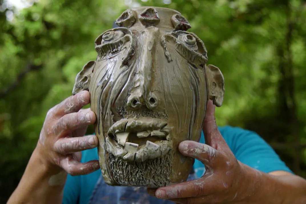 A man holds up a face jug, blocking his own face.