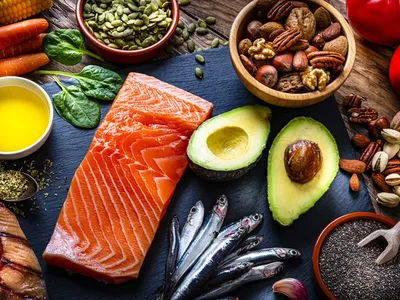Image shows a variety of foods including a piece of salmon, avocados, sardines and two small bowls of nuts and seeds