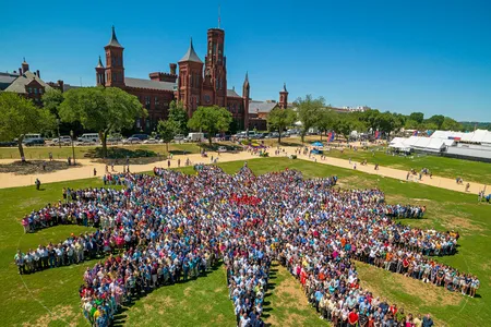 Thousands of people assemble on the green grass of the National Mall to form a sunburst shape, the Smithsonian logo. The red-brick Smithsonian Castle rises in the background.