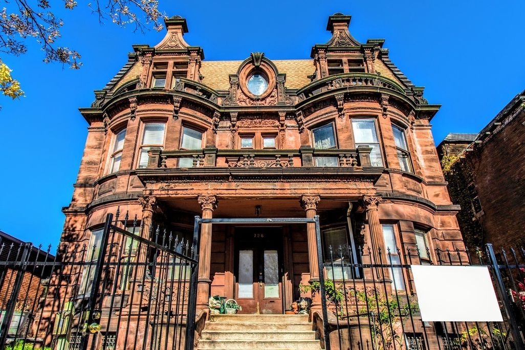 A front view of an ornate brownstone mansion, with columns, a circular window in the center, turret-like shapes on either side and a peaked roof