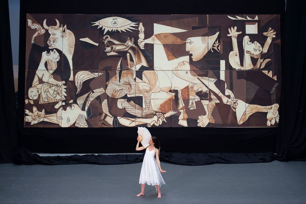 A person in a white flowy dress and holding a white fan dances in front of the enormous chocolate replica of Picasso's Guernica