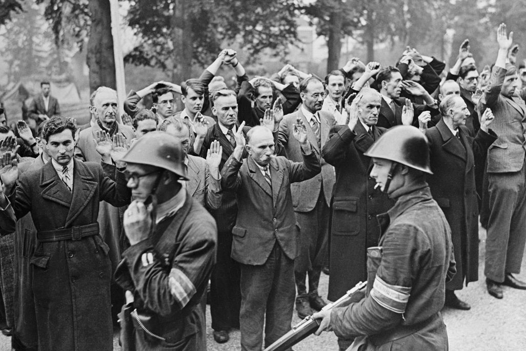Black and white view of group of men holding their hands in the air with men in uniform in the foreground